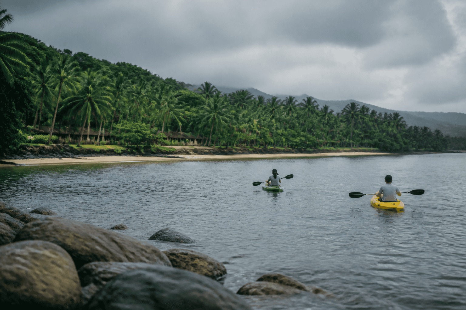Two paddlers move across calm grey-blue water beside a rain-washed tropical coast under overcast sky.