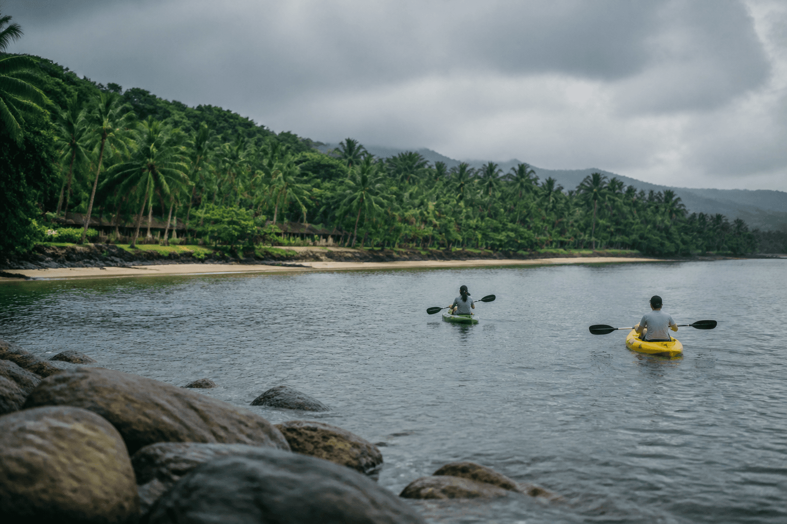 Two paddlers move across calm grey-blue water beside a rain-washed tropical coast under overcast sky.