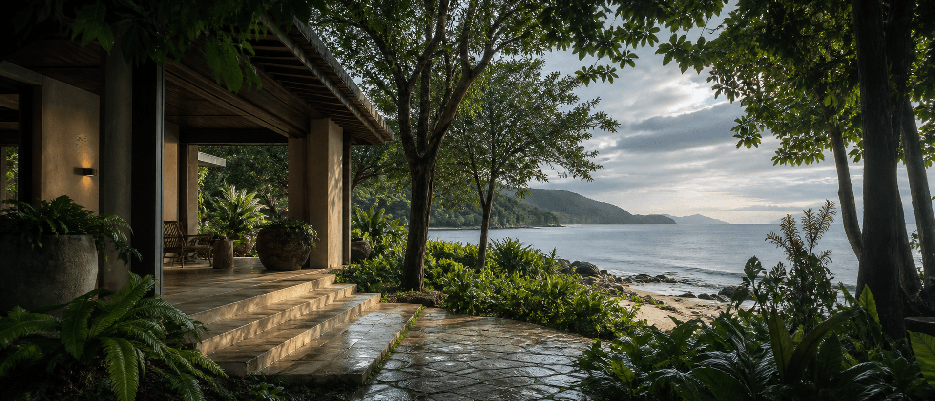 A shaded, single-level coastal pavilion opens through a rain-washed grove toward a broad grey-blue horizon.
